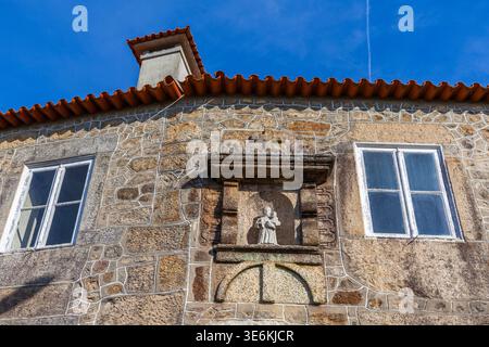 Eine traditionelle Fassade aus Granitstein verfügt über weiß gerahmte Fenster und eine zentrale religiöse Nische mit einer Statue unter einem Terrakottadach Stockfoto