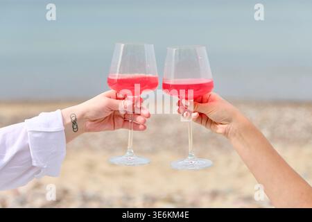 Nahaufnahme von zwei weiblichen Händen, die beim Strandpicknick pinkfarbene Weingläser klirren. Ruhiger Meereshintergrund und sanfte natürliche Beleuchtung sorgen für eine warme, freundliche Atmosphäre Stockfoto