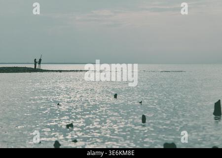 Ein einsamer Fischer steht an der felsigen Küste, die in ruhiges Wasser unter bewölktem Himmel mit sanfter, gedämpfter Beleuchtung mündet. Friedliche Küstenszene vermittelt s Stockfoto