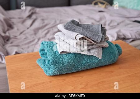 Weiches, kuscheliges türkisfarbenes Handtuch mit kleineren, ordentlich gefalteten grauen Handtüchern, die oben auf polierter Holzoberfläche im Schlafzimmer mit gedämpfter Oberfläche platziert werden Stockfoto