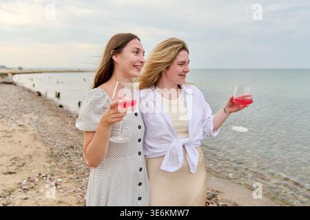Zwei junge Frauen, die lächeln und Gläser mit rosafarbenem Wein halten, stehen am Rocky Beach in der Nähe des ruhigen Meeres unter weichem Tageslicht. Ihre fröhlichen Ausdrücke heben Wa hervor Stockfoto