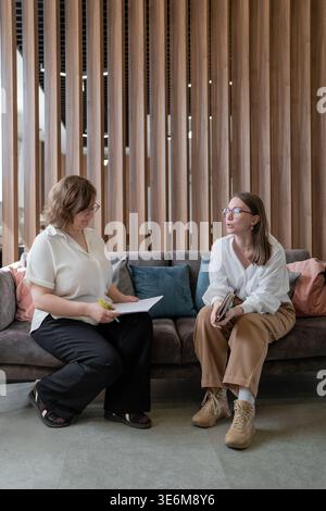 Frauen, die nebeneinander auf der Couch sitzen und in einem modernen Büro mit vertikaler Holzeinrichtung geschäftlich sprechen. Business, Talk, Couch, Frauen, modern, Einrichtung. Stockfoto