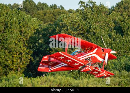 Leuchtend rotes Kunstflugzeug fliegt während einer Outdoor-Airshow tief über grüne Bäume und zeigt Vintage-Styling, präzises Fliegen und dynamische Bewegung ag Stockfoto