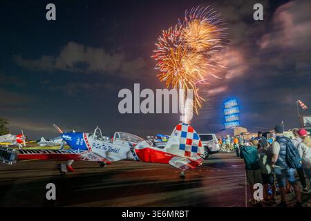 Die Zuschauer beobachten ein patriotisches rot-weiß-blaues Kunstflugzeug, das auf dem Asphalt geparkt ist, während in einer lebhaften Nacht ein farbenfrohes Feuerwerk über dem Boden explodiert Stockfoto