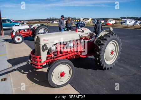 Roter und cremefarbener Ford-Traktor, der auf einem kleinen Flughafenparkplatz mit leichten Flugzeugen und Zuschauern in der Nähe geparkt wurde. Heller, sonniger Himmel, ländlicher Flugplatz Stockfoto