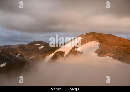 Berggrat mit schmelzenden Schneeflächen, die durch dicken Morgennebel auftauchen. Dramatische Wolken und Nebel sorgen für stimmungsvolle Atmosphäre im Hochland Stockfoto