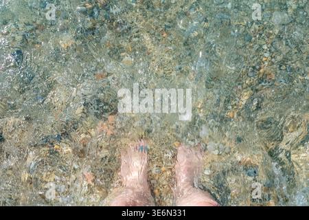 Ein Blick von oben auf klares Meerwasser und weißen Schaum, der über die Füße einer Person am Sandstrand gespült wird. Erfrischende Szene eines Sommerurlaubs am Meer Stockfoto