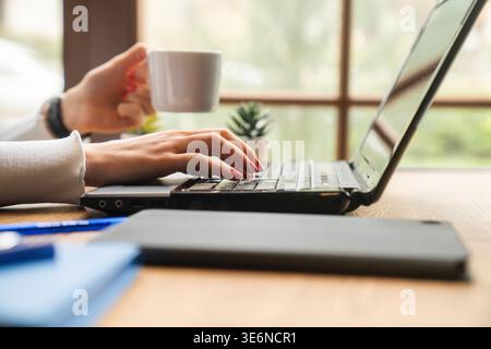 Eine Frau mit rotem nagellack hält eine Kaffeetasse, während sie an einem sonnendurchfluteten Arbeitsplatz auf einem Laptop schreibt. Dies bedeutet freiberufliches Arbeiten, Fernarbeit, ein digitales Nein Stockfoto
