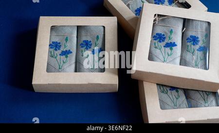 Leinenservietten mit zarter blauer Kornblumenstickerei in umweltfreundlichen Papierkartons, selektiver Fokus. Elegantes, besticktes Textilezubehör Stockfoto