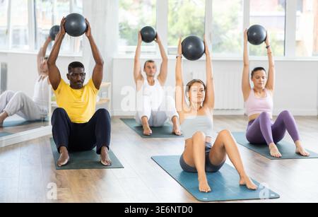 Pilates-Klasse in multiethnischen Gruppen - Männer und Frauen halten den Ball über den Kopf und beugen sich Stockfoto