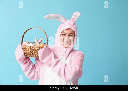 Lustige Seniorin im Osterhasen-Kostüm, der Korb mit Ostereiern auf blauem Hintergrund hält Stockfoto