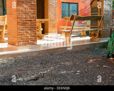 Holzbank auf einer Veranda neben Ziegelsäulen. Sonnenlicht wirft Schatten auf Kiesboden. Rustikale Sitzgelegenheiten im Freien laden zur ruhigen Entspannung ein. Szenenanzug Stockfoto