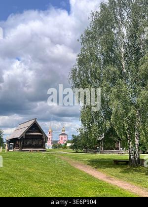 Historische Holzgebäude im Museum für Holzarchitektur in Suzdal, Russland, mit sichtbaren Kirchtürmen im Hintergrund. Stockfoto