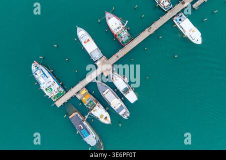 Eine Hochwinkeldrohne zeigt verschiedene Boote, einschließlich Fischerboote und Yachten, die an Piers in einem klaren, türkisfarbenen Hafen angedockt sind. Stockfoto