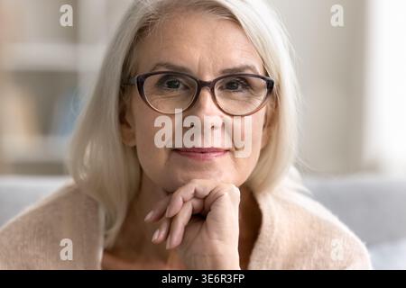 Eine ältere Frau trägt eine Brille, liegt auf dem Sofa, legt das Kinn auf die Hand, starrt in die Kamera. Porträt einer gepflegten, schönen, reiferen Frau, Stockfoto