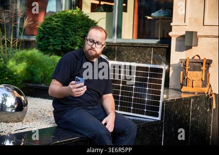 Bärtiger Mann, der ein Smartphone neben dem Photovoltaik-Sonnenkollektor benutzt. Integration nachhaltiger erneuerbarer Energien in den Alltag, demonstrieren die praktische Nutzung von Solarstrom für Ladegeräte. Stockfoto