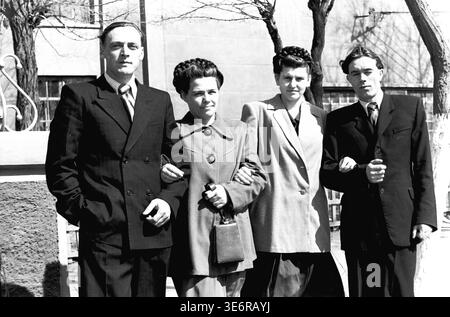 Sloviansk, 1950. Außerhalb der Anker- und Isolieranlage (AIZ). Vier junge sowjetische Fabrikkollegen stehen stolz Arm in Arm und posieren für ein freundliches Gruppenporträt an einem sonnigen Tag vor einer festlichen Massendemonstration. Die Männer tragen scharfe dunkle Anzüge und die Frauen tragen elegante Mäntel aus der Mitte des Jahrhunderts mit wunderschönen geflochtenen Frisuren. Dieses äußerst authentische Schwarzweiß-Archivfoto zeigt die Freundschaft am Arbeitsplatz, die Jugendkultur und das Erbe des friedlichen Donbass perfekt. Stockfoto