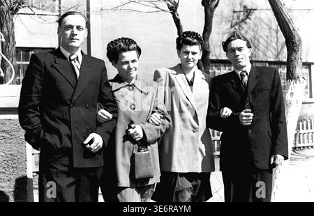 Sloviansk, 1950. Außerhalb der Anker- und Isolieranlage (AIZ). Vier junge sowjetische Fabrikkollegen stehen stolz Arm in Arm und posieren für ein freundliches Gruppenporträt an einem sonnigen Tag vor einer festlichen Massendemonstration. Die Männer tragen scharfe dunkle Anzüge und die Frauen tragen elegante Mäntel aus der Mitte des Jahrhunderts mit wunderschönen geflochtenen Frisuren. Dieses äußerst authentische Schwarzweiß-Archivfoto zeigt die Freundschaft am Arbeitsplatz, die Jugendkultur und das Erbe des friedlichen Donbass perfekt. Stockfoto