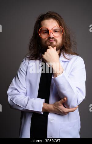 Studio-Porträt eines bärtigen jungen Mannes mit langen Haaren in weißem Laborkittel und übergroßer oranger Brille, der sein Kinn nachdenklich hält. Kreativ A Stockfoto
