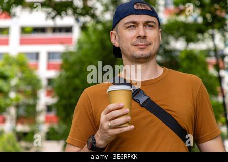 Mann in lässigen Kleidern, der eine Kaffeetasse hält und im Stadtpark steht. Authentisches Lifestyle-Porträt von männlichen Touristen oder Pendlern während der morgendlichen Pause Stockfoto