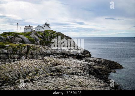 Weiße Leuchtturmgebäude auf felsigen Klippen mit Blick auf das ruhige Meer unter bewölktem Himmel in abgelegener Küstenlandschaft. Stockfoto