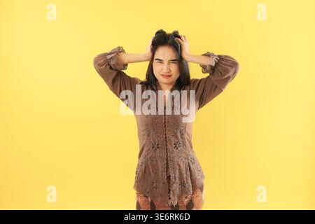 Der wütende oder verrückte Ausdruck und die Geste der indonesischen Frau in Brown Kebaya und Batik, isoliert auf gelbem Hintergrund. Asiatische Frau kratzt sich an irrit Stockfoto