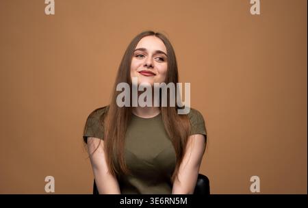 Junge Frau mit langen braunen Haaren posiert vor beigefarbenem Hintergrund, lächelt und schaut nach oben zur Seite Stockfoto