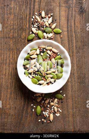 Mischung aus Samen in weißer Schüssel auf hölzernem Hintergrund, Draufsicht. Gesunde Ernährung Inhaltsstoffe Textur. Kürbiskerne, schwarzes und weißes Sesam, Sonnenblumenkerne Stockfoto