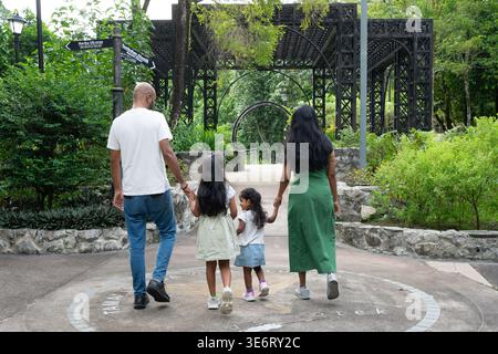 Eltern mit zwei Töchtern gehen Hand in Hand auf einem Pfad in einem üppigen grünen Park Stockfoto