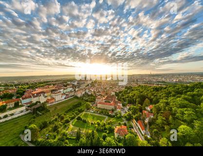 Die Sonne geht über Prag auf und wirft goldenes Licht über die Dächer und die baumbestandenen Hügel der Stadt. Die Moldau schimmert in der Ferne, während historische Gebäude sich am Himmel abheben. Stockfoto