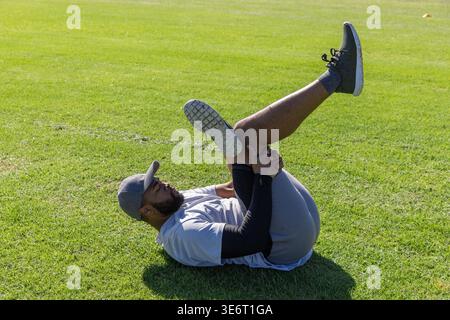 Afroamerikaner, der auf dem Rücken liegt und das Bein über die Brust auf dem Sportplatz hält, mit weißen Markierungen Stockfoto