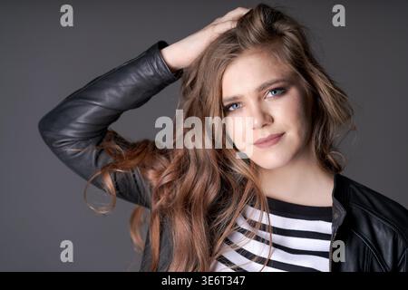 Eine Frau mit langen braunen Haaren posiert für ein Foto. Sie trägt ein schwarz-weiß gestreiftes Hemd und eine Lederjacke. Image hat eine coole und scharfe Atmosphäre mit langen Haaren der Frau Stockfoto