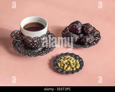 Eine Tasse Kaffee mit Kardamom ruht neben getrockneten Datteln Früchten Dunkle Ajwa wagt es in einer herzförmigen Schüssel zu sitzen. Grüne Samen leuchten in einer kleinen Schüssel. Rosa Oberfläche hält alles ruhig. Gemütliche, warme Atmosphäre für Frühstück oder Tee Stockfoto