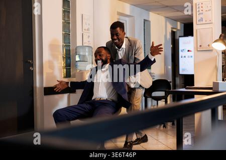 afroamerikanische Arbeiter, die im modernen Startup-Büro Sportwettbewerbe veranstalten, eine Pause von der Arbeit machen und Spaß haben. Fröhliche Geschäftsleute in formellen Anzügen, die bis spät in die Nacht einen Schreibtischstuhl tragen Stockfoto