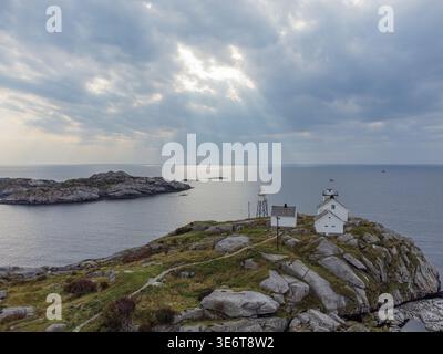 Aus der Vogelperspektive eines kleinen weißen Leuchtturms, der stolz auf einem Felsvorsprung steht, geküsst vom Sonnenlicht, das durch dramatische Wolken filtert, Henningsvær, Nordla Stockfoto