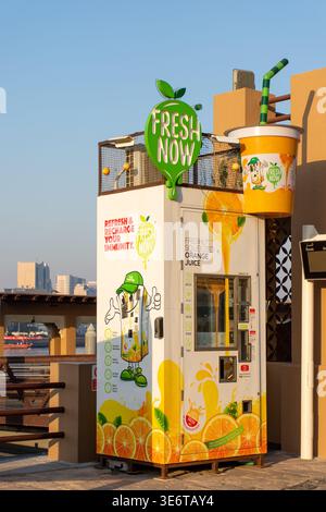 Automatisierter und frisch gepresster Orangensaft-Automat mit farbenfroher Branding an einer sonnigen Uferpromenade in Dubai, VAE. Stockfoto