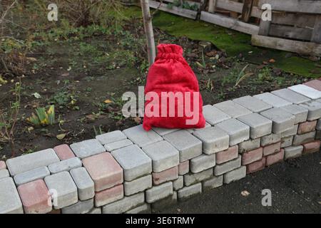 Eine rote Tasche wird auf einem Steinweg in einem Garten platziert. Die Umgebung ist von Pflanzen und Bäumen umgeben. Das Licht ist weich und zeigt den späten Nachmittag an. Die Blätter sind auf dem Boden verstreut. Stockfoto