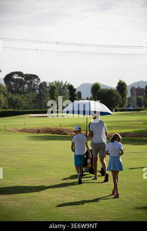Vater als Caddy, der mit Kindern auf dem Golfplatz mit Sonnenschirm an warmen sonnigen Tagen in Italien spaziert Stockfoto
