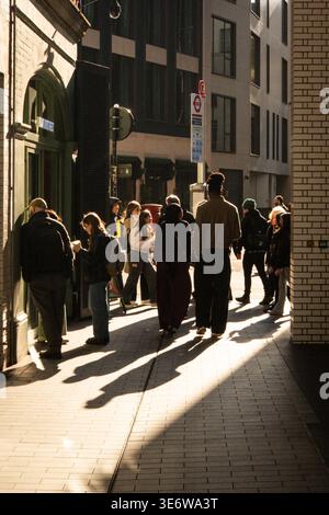 Fußgänger laufen durch eine enge Londoner Straße bei niedriger Wintersonne und werfen lange Schatten über den Bürgersteig. Stockfoto