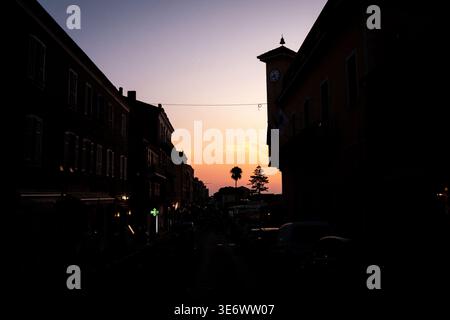 In Propriano, Korsika, Frankreich, Augus, ist eine Straße mit dunklen Gebäudesilhouetten, geparkten Autos, einem Uhrenturm und einem schwindenden Sonnenuntergang zu sehen Stockfoto