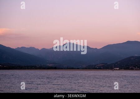 Meerwasser erstreckt sich über den Vordergrund unter einem rosafarbenen Abendhimmel mit dunklen Bergsilhouetten am Horizont in Propriano, Korsika, Frankreich, Augu Stockfoto