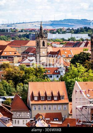 Historisches europäisches Stadtbild mit roten Ziegeldächern, zentralem Glockenturm aus Stein, grünen Bäumen im Vordergrund und sanften Hügeln mit Windturbinen im Hintergrund Stockfoto