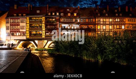 Nächtliche Flussszene mit beleuchteten Fachwerkgebäuden, die sich im Wasser spiegeln, bogenförmige Steinbrücke, üppige Ufervegetation und stimmungsvoll bewölktem Himmel. Stockfoto