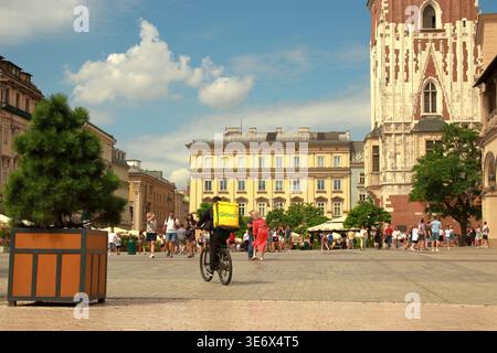 Glovo-Kurier radelt durch einen belebten Platz in Krakau mit historischen Gebäuden, Fußgängern und Cafés im Hintergrund. Stockfoto