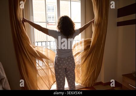 Eine Frau im Pyjama mit Sternenmuster öffnet transparente Vorhänge, um warmes Morgensonnenlicht in einem gemütlichen Schlafzimmer willkommen zu heißen, was das Aufwachen, die ruhige Routine, die Erneuerung und das Leben suggeriert Stockfoto