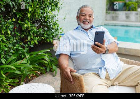 Ranghoher afroamerikanischer Mann, der sich im Korbstuhl auf dem Pooldeck entspannt, Telefon hält und eine Uhr trägt Stockfoto