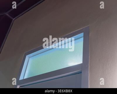 Niedriger Winkel auf ein beleuchtetes rechteckiges Fenster mit geripptem Milchglas, das nachts in eine glatte beige Außenwand des Gebäudes eingefasst ist. Die Szene repräsentiert Stockfoto