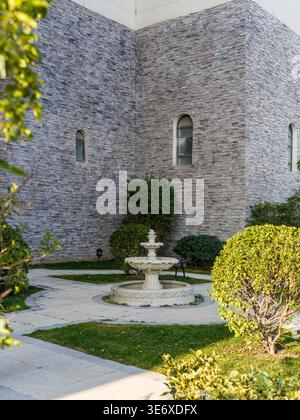 Friedlicher geschlossener Innenhof, umgeben von strukturierten grauen Steinmauern, mit einem kleinen klassisch gestuften Brunnen, grünen gepflegten Büschen und p Stockfoto
