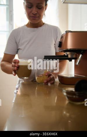 Selektive Hände auf den Händen einer verschwommenen jungen Frau in der Küche, die neben ihrer Kaffeemaschine steht und ein Glas frisch zubereiteten Cappuccino hält. Stockfoto
