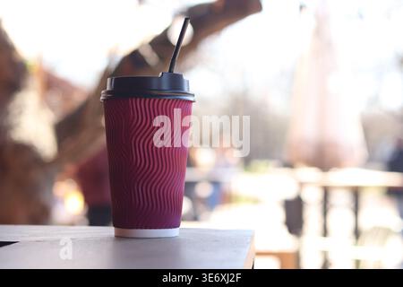 Roter Papierbecher mit gewellter Oberfläche und schwarzem Deckel und Strohhalm, der auf einem Holztisch vor einem verschwommenen Hintergrund eines Sommercafés steht. Zum Mitnehmen Stockfoto